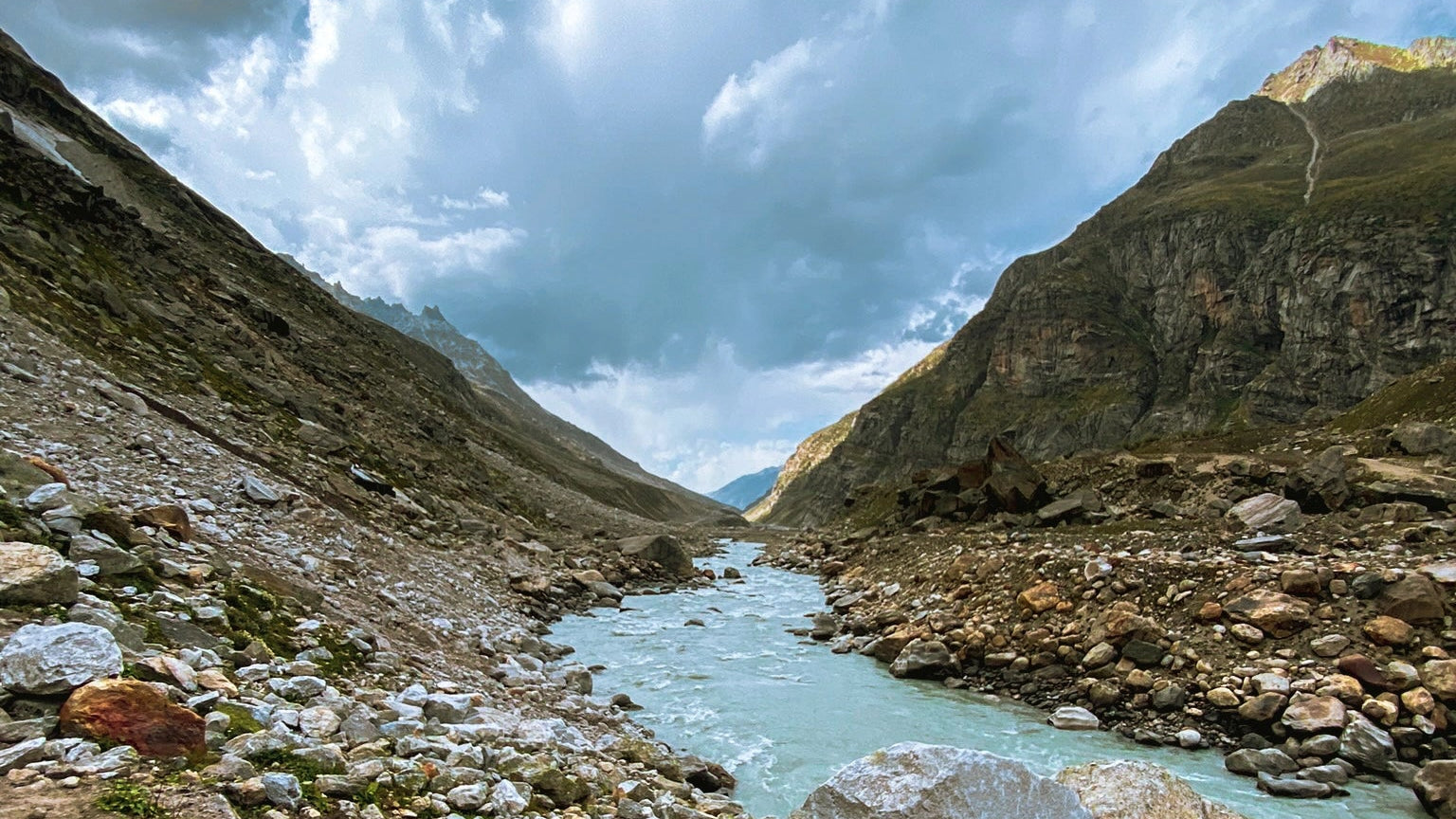 Mountainous landscape with a river flowing through rocky terrain under a cloudy sky.
