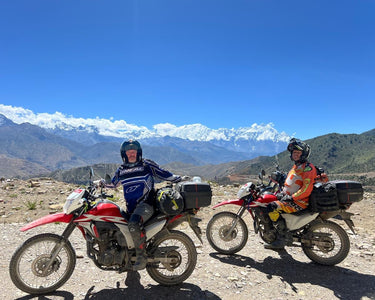 Two riders on dual-sport motorcycles on a mountain track with snow-capped Himalayan peaks behind