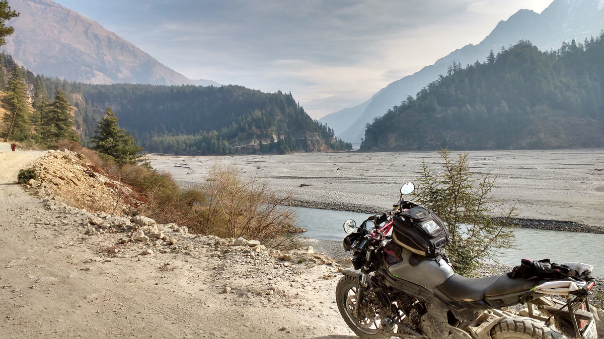 Loaded motorcycle parked on a dirt track beside a wide braided riverbed in a forested valley under hazy skies