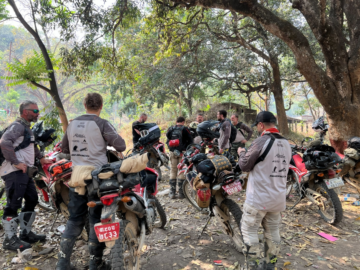 Riders gathered under large trees beside parked motorcycles