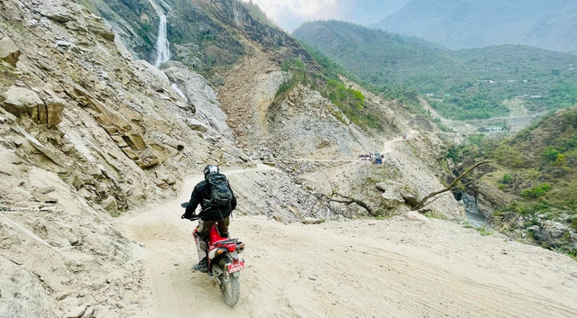 Rider on a rough mountain road cut into a rocky hillside, with a waterfall, green hills and vehicles further along the road